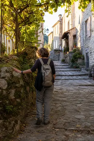 Quiet alleyway in an Ardèche village, a woman stands contemplatively by a mossy stone wall in gentle evening light.
