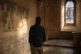 Middle-aged artisan, seen from behind, studies old frescoes inside a sunlit stone chapel in Ardèche at golden hour, with soft natural light and serene atmosphere.