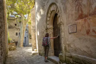 Middle-aged hiker from behind touching an old Romanesque chapel doorway in a sunlit stone alley, village atmosphere, soft evening light.