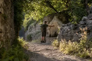Shaded village path with stone walls, lone hiker touching old fountain, golden afternoon light, quiet and authentic mood