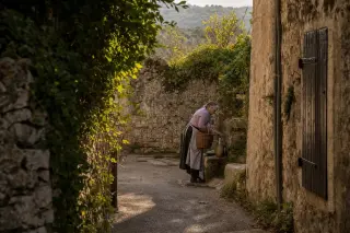 Elderly woman in a stone alley of Labeaume filling a jug at an ancient fountain, sunlight on the walls, scene viewed through climbing vines for an authentic, intimate atmosphere.