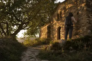 Stone footpath through wild grass leading to a small Romanesque chapel, with a hiker gently touching the mossy wall in late afternoon light.