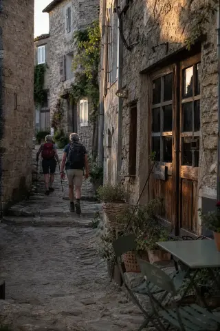 Two hikers climb an old stone alley in a tranquil hilltop village at dusk, soft golden light highlighting worn steps and textured walls, empty café chairs suggesting everyday life.