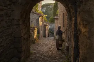Elderly woman with basket touching ancient village wall in warm evening light, narrow medieval lane in Balazuc