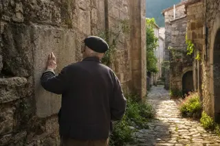 Elderly man in rustic clothing calmly touches an ancient stone chapel wall in a sunlit, narrow village alley, evoking a quiet moment of connection with Ardèche heritage.