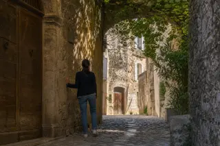 Person standing in a shaded medieval alleyway in Viviers, gently touching a stone wall, bathed in soft late afternoon light with vine shadows nearby.